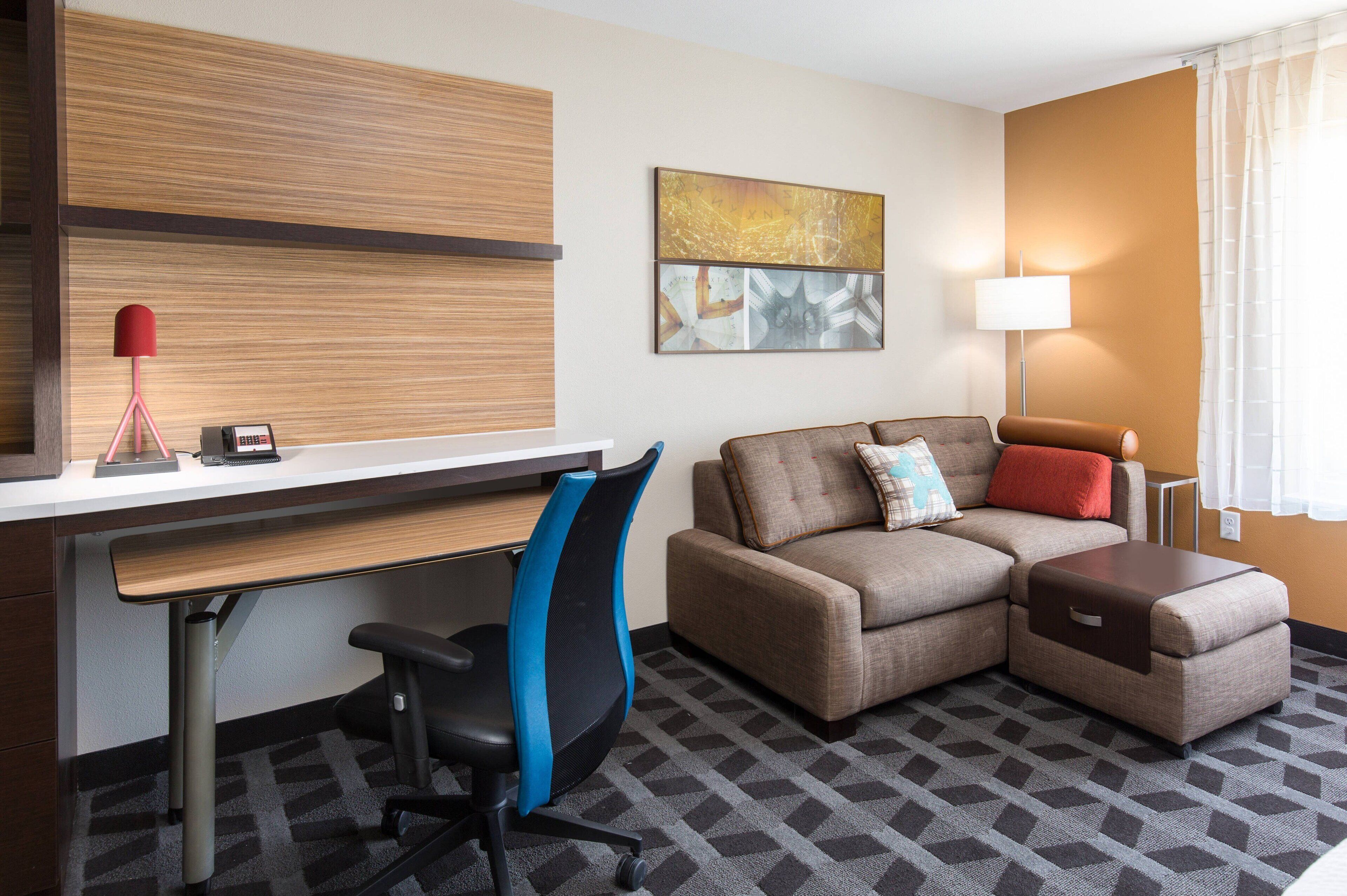 A modern hotel room with a desk, office chair, sofa, ottoman, and a lamp.
