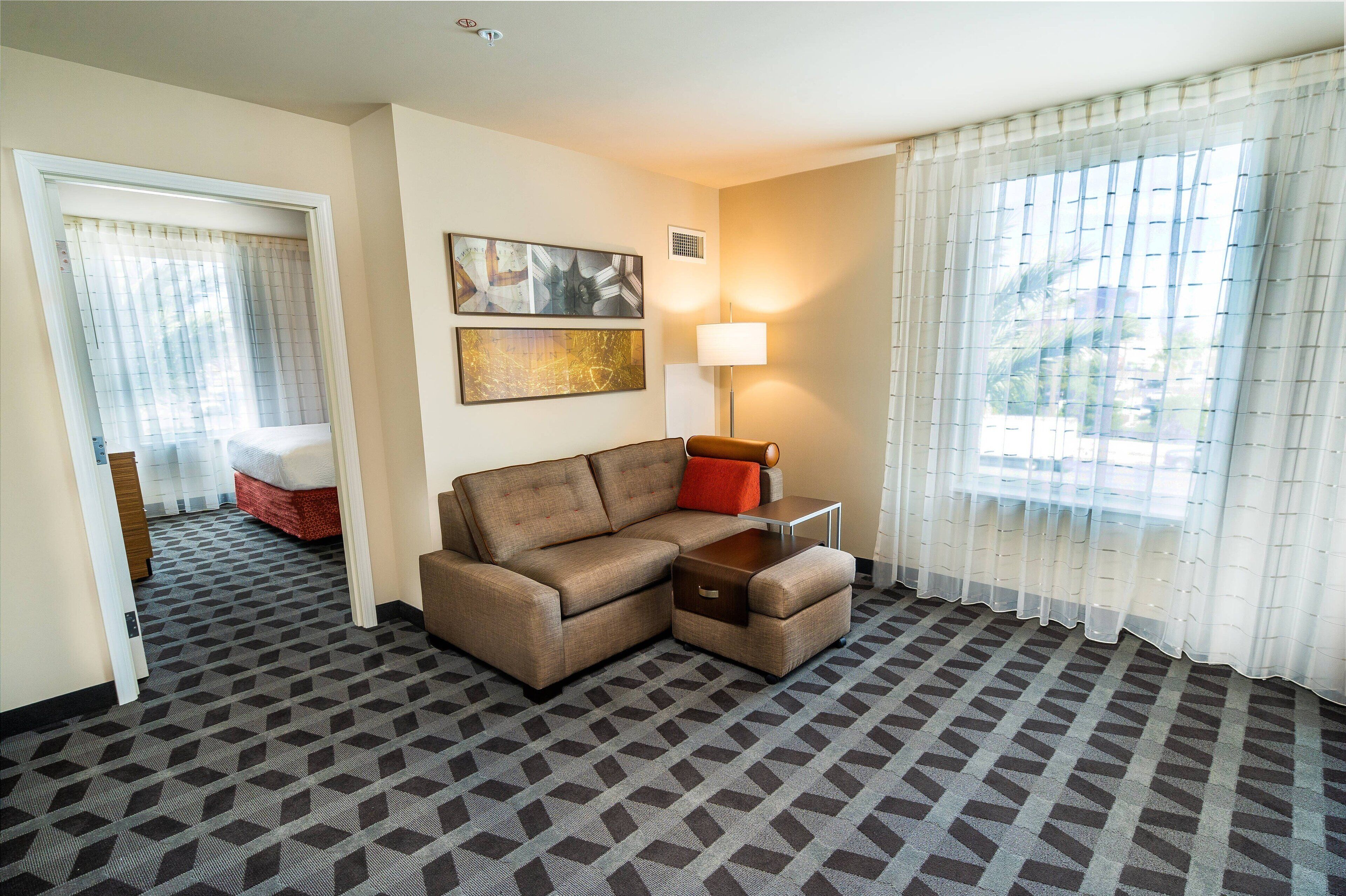 A hotel room with a sectional sofa, a small table, a lamp, and a view of the outdoors through sheer curtains.