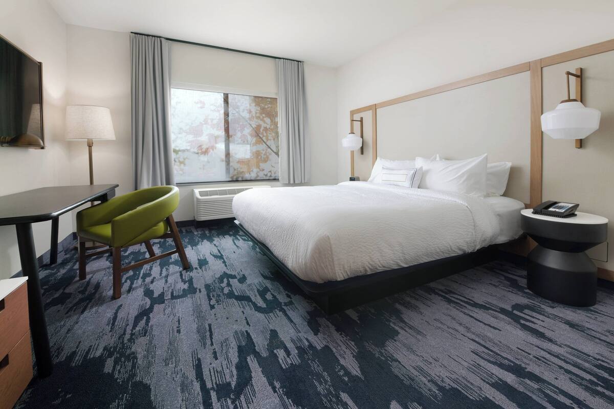 A hotel room with a large bed, a desk, a chair, a bedside table with a lamp, and a window with curtains.