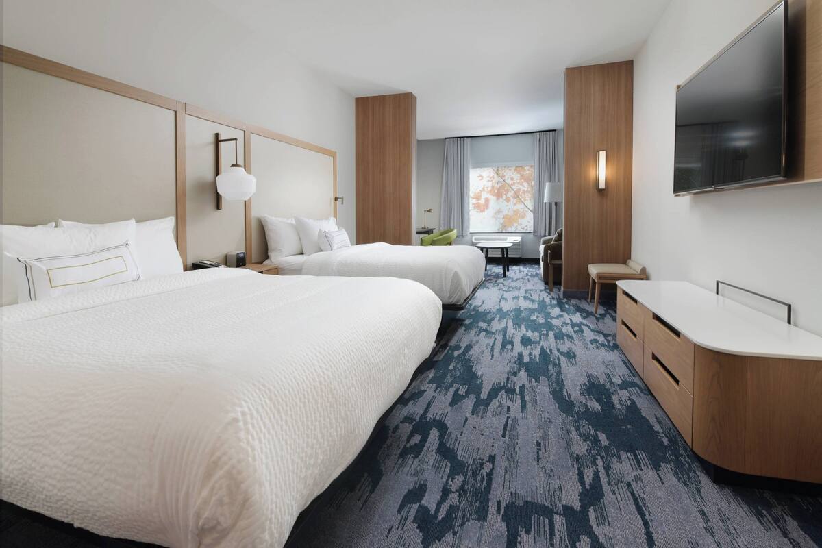 A hotel room with two large beds, a desk, a chair, a TV, and a large window.