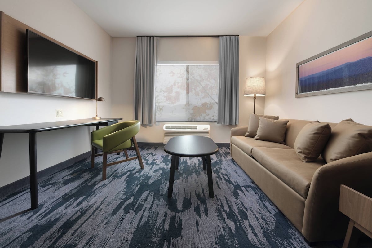 A hotel room with a flat-screen TV, a sofa, a small table, a green chair, and a large window with curtains.