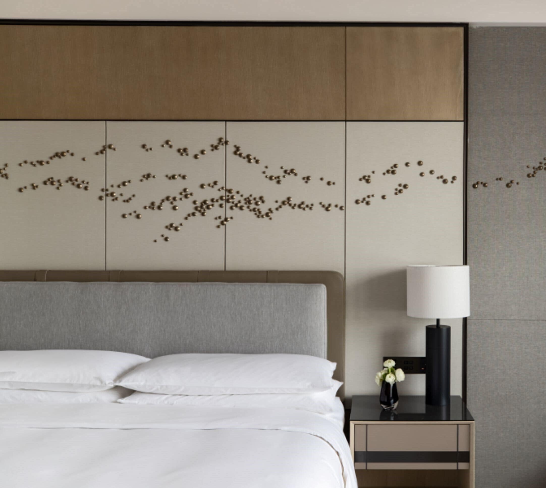 Quarto de hotel com cama, mesa de cabeceira com luminária e painel decorativo na parede.