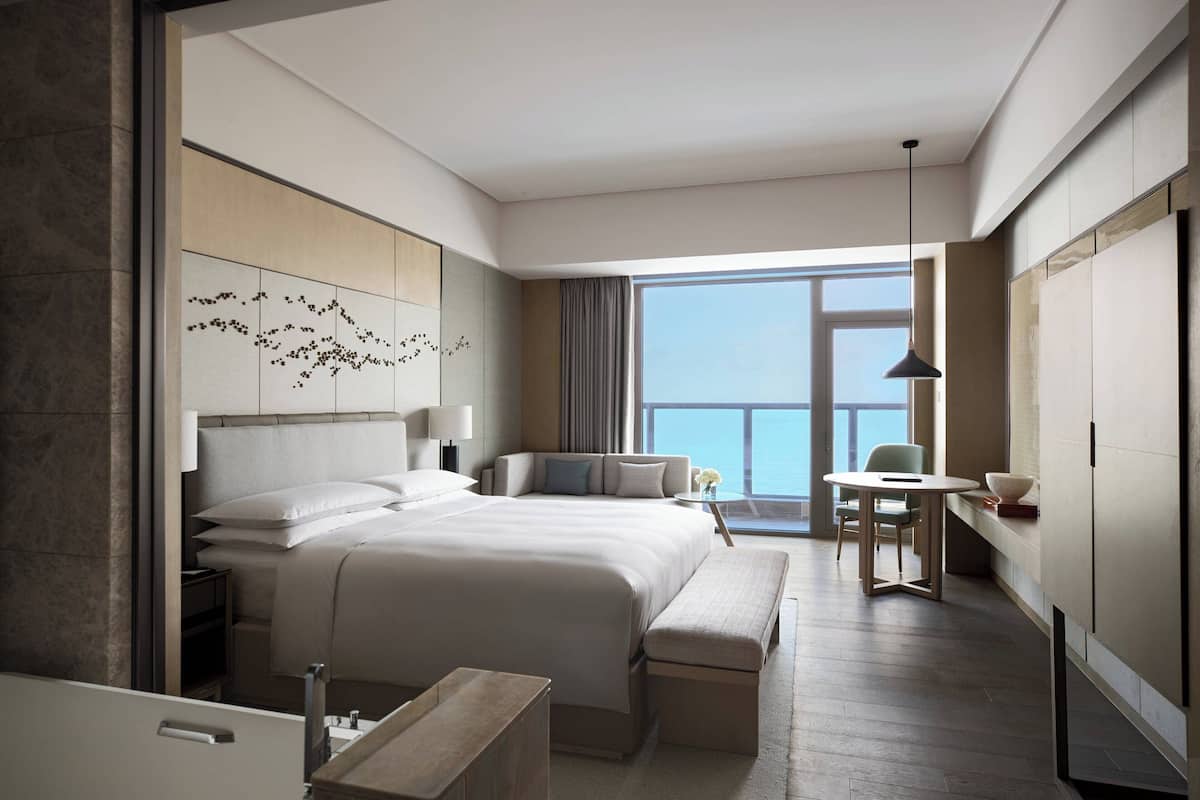 A modern hotel room with a large bed, a sitting area with a table and chairs, a small balcony, and a view of the sky.