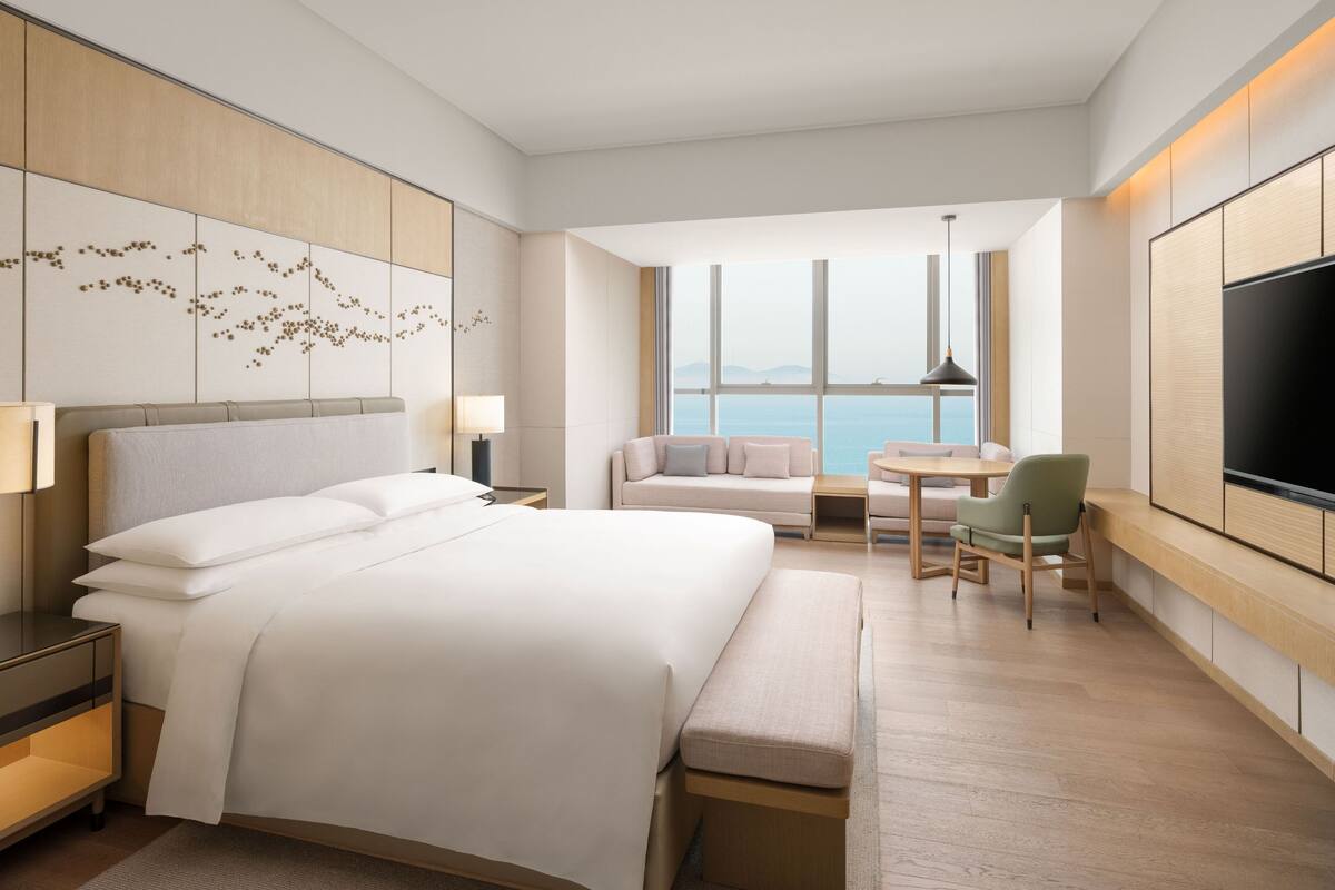 A modern hotel room with a large bed, a seating area with a table and chairs, a TV, and a view of the sea through large windows.