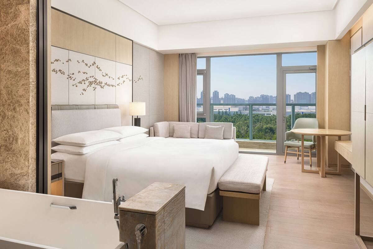 A modern hotel room with a large bed, a sitting area, a small table, and a view of the city.