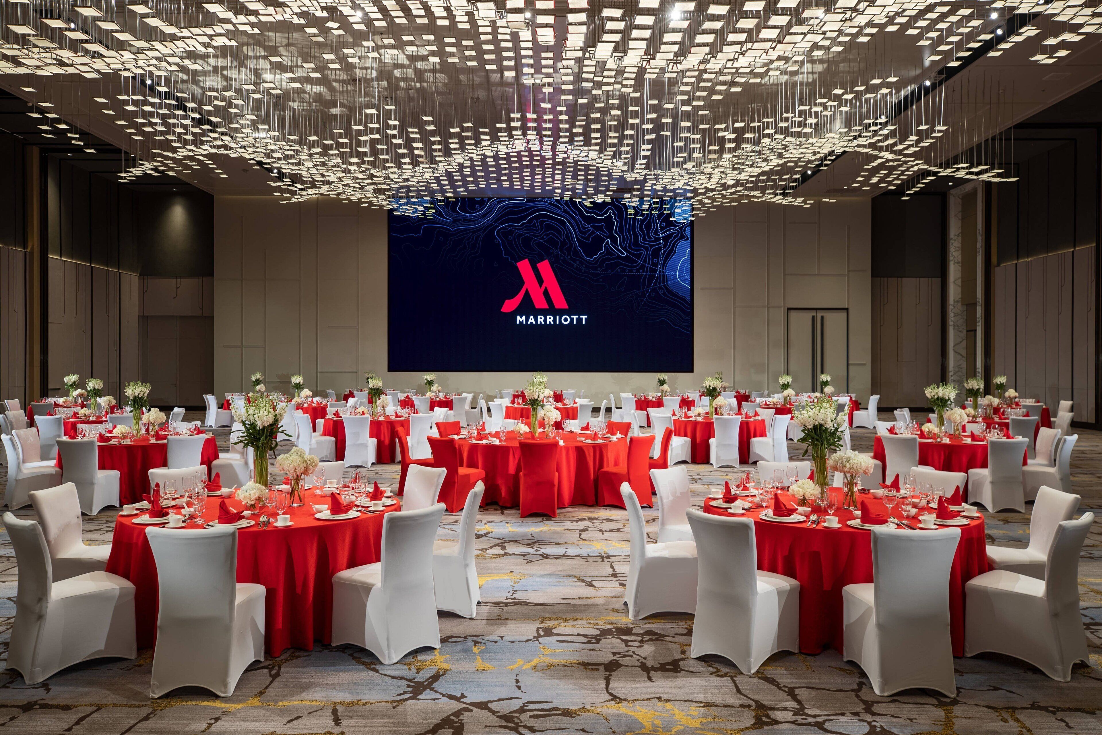 Una vasta sala conferenze con tavoli apparecchiati per un evento formale, con un logo Marriott ben visibile sullo schermo.
