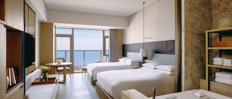 Et moderne hotelværelse med to senge, et opholdsområde med et bord og stole, et stort vindue med udsigt, et badeværelse med badekar og en hylde med håndklæder.