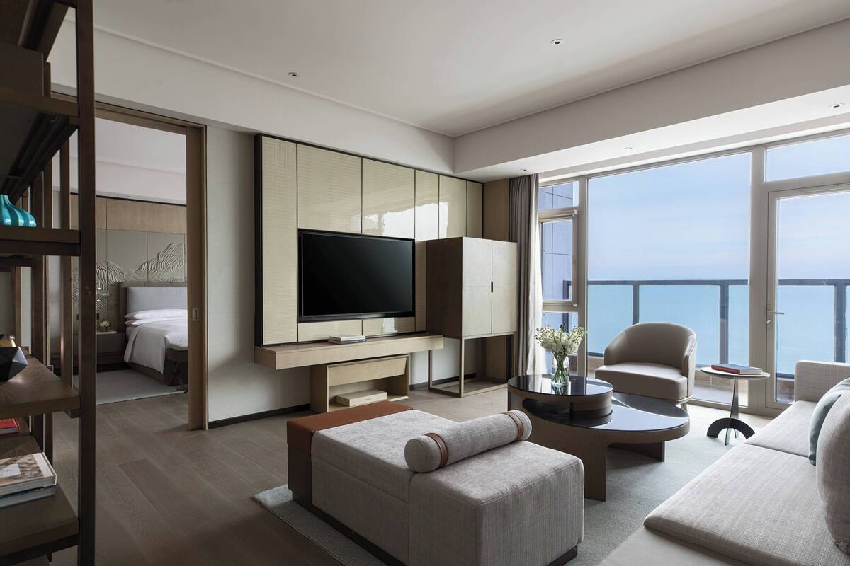 A modern hotel room with a large TV, comfortable seating, a balcony with a view, and a minimalist design.