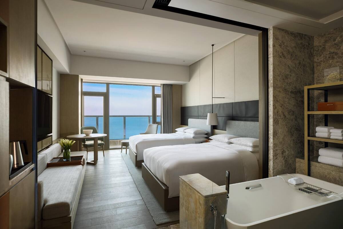 A hotel room with two beds, a sitting area, a large window with a view, a bathtub, and a shelf with towels.