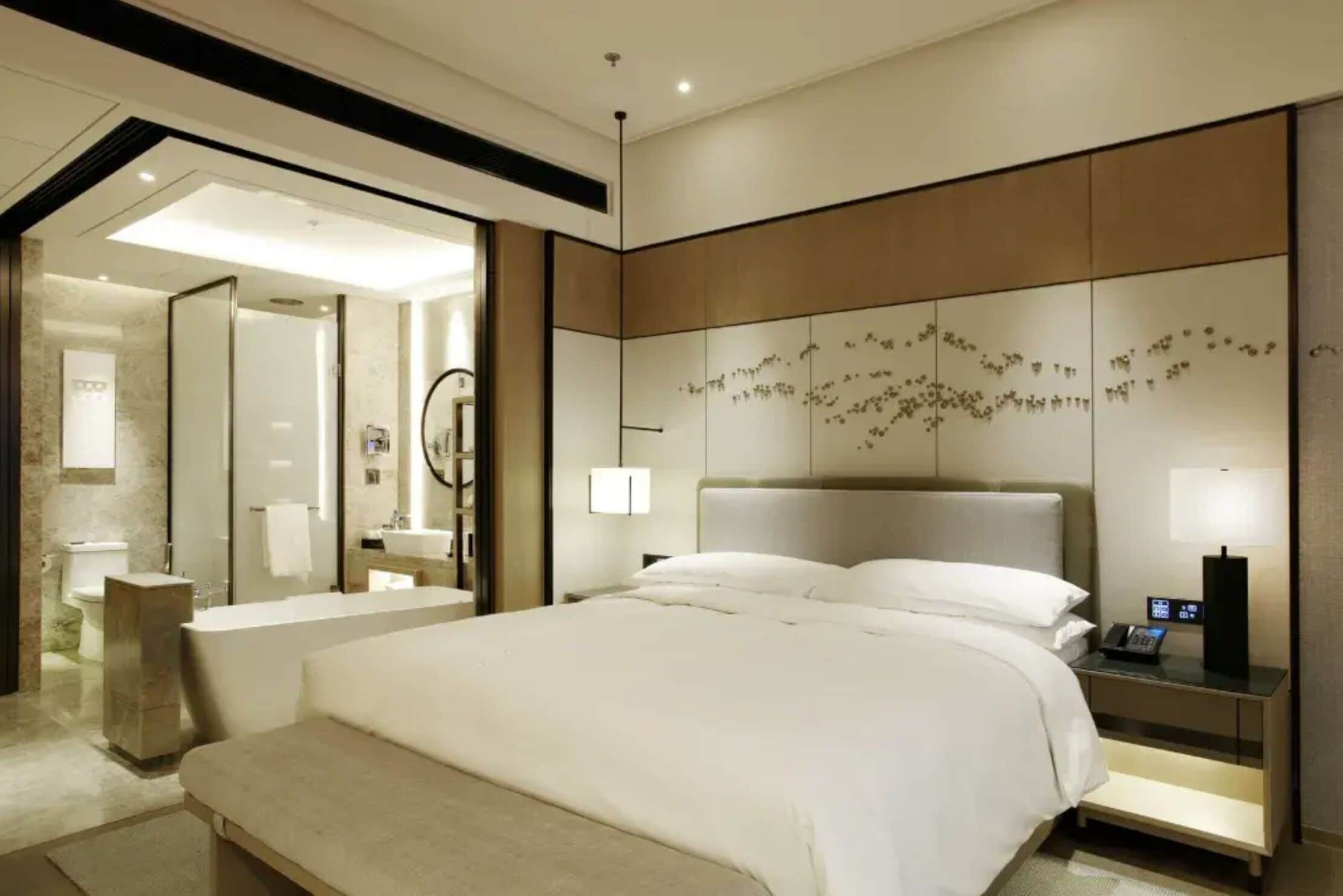 A modern hotel room with a large bed, a bedside table with a phone, a wall-mounted artwork, and a bathroom visible through an open door.