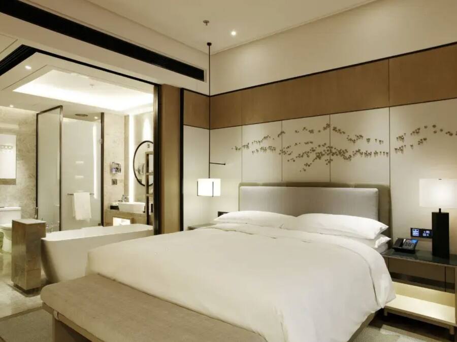 A modern hotel room with a large bed, a bedside table with a phone, a wall-mounted artwork, and a bathroom visible through an open door.