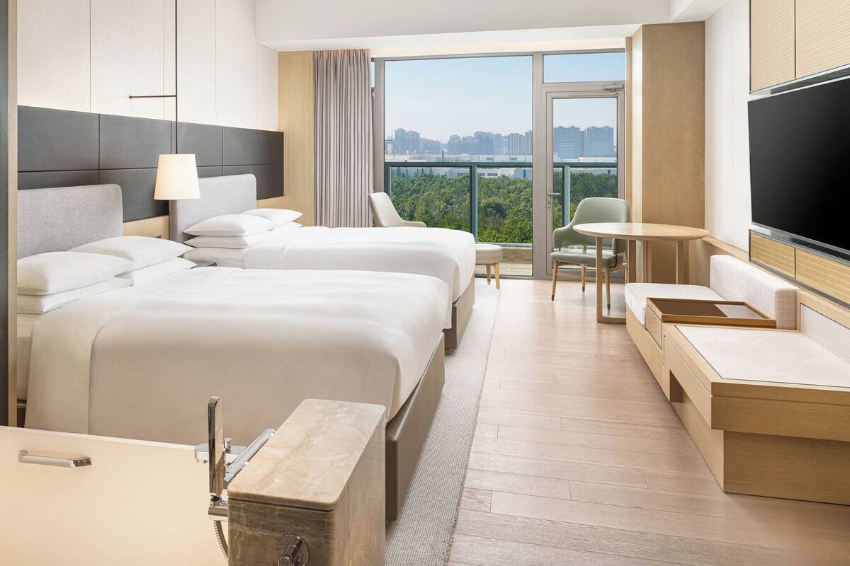 A hotel room with a large bed, a desk, a chair, a TV, and a balcony with a city view.