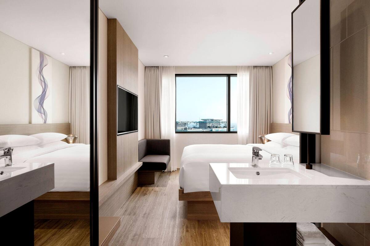 A modern hotel room with two beds, a large window, a TV, and a bathroom with a large sink.