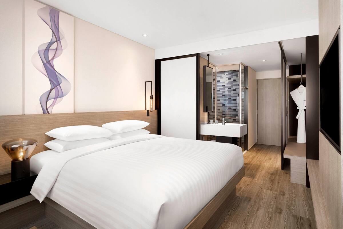A modern hotel room with a large bed, a built-in wardrobe, a bathroom with a tiled shower, and a wall-mounted artwork.