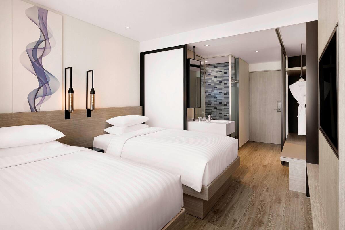 A modern hotel room with two beds, a large mirror, a bathroom with a tiled backsplash, and a robe hanging on a hook.