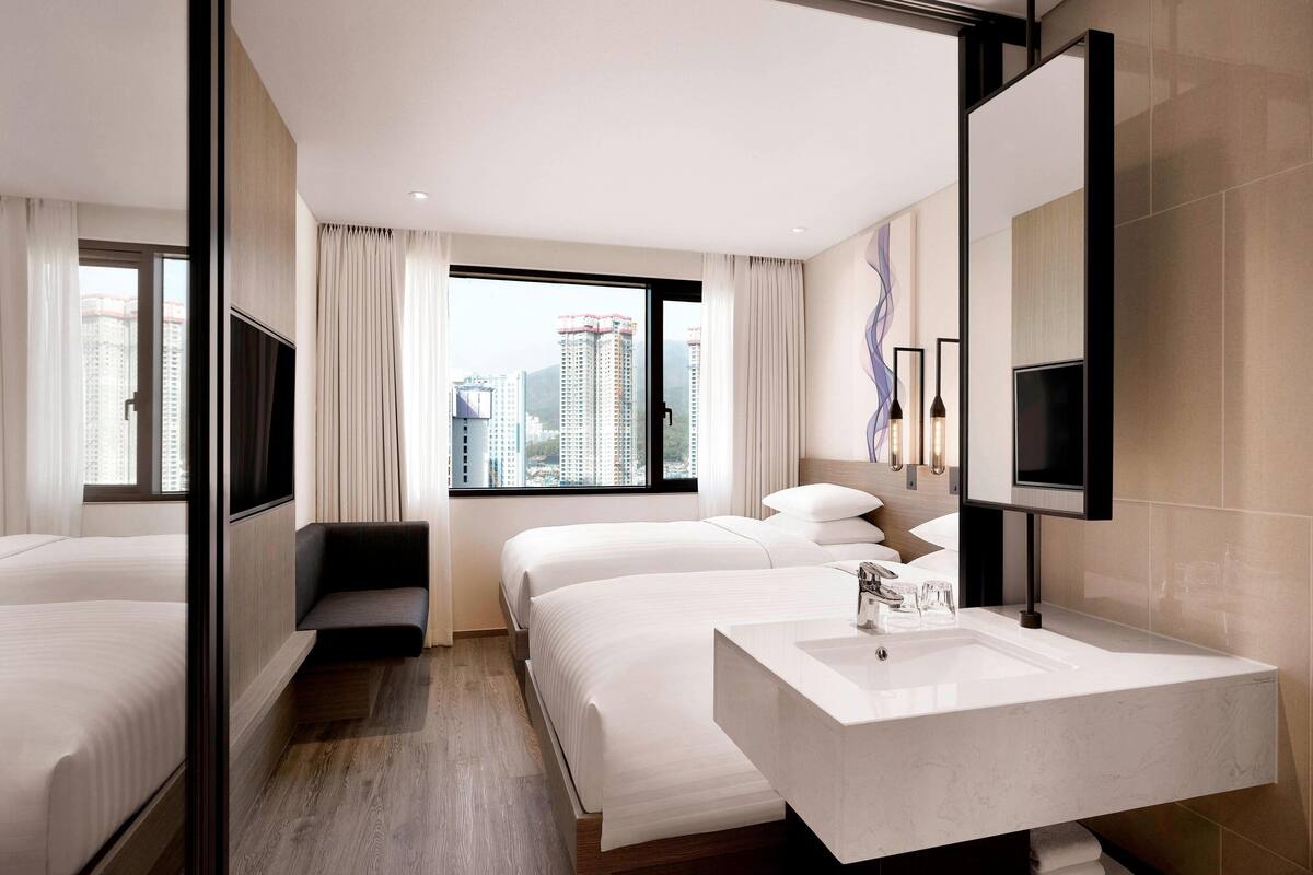 A hotel room with two beds, a large window with curtains, a modern bathroom with a vanity and sink, and a city view.