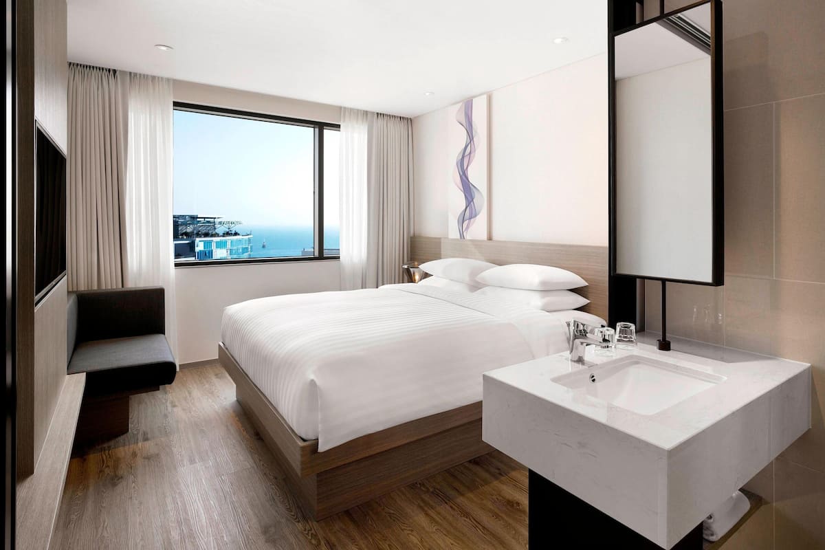 A modern hotel room with a large bed, a built-in bench, a large window with a view, and a modern bathroom with a large sink and mirror.