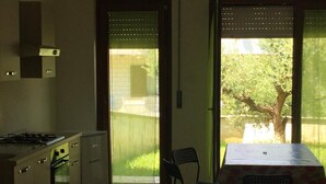 Cocina con estufa a gas, horno y refrigerador. Cuenta con un comedor con mesa y sillas, y vistas a un espacio exterior verde a través de una puerta.