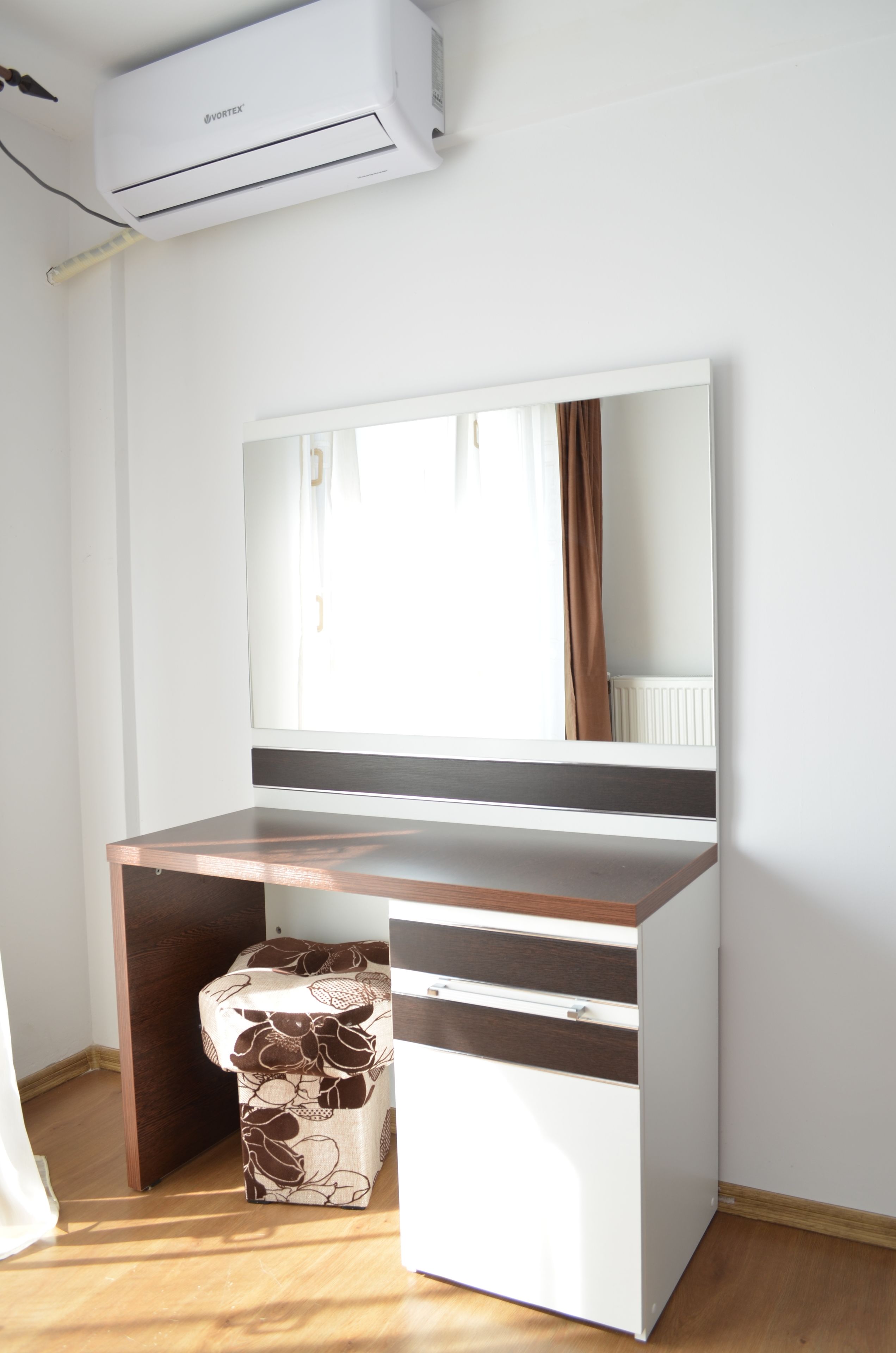 Una habitación compacta con un escritorio de madera, una silla blanca con estampado floral, un espejo grande y un equipo de aire acondicionado instalado en la pared.