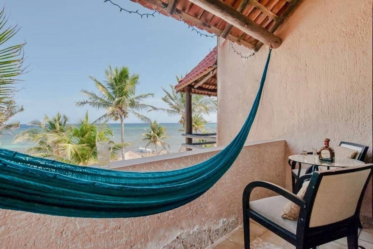 Un balcón con una hamaca, una mesa con sillas y vistas al océano y palmeras.