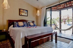 A bedroom with a bed, a bench, a nightstand, a painting, and a view of the outdoors. - Hammock Heaven (Akumal)