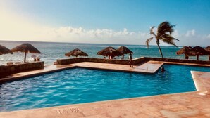 Zona de piscina con vista al océano, sombrillas de paja y palmeras.