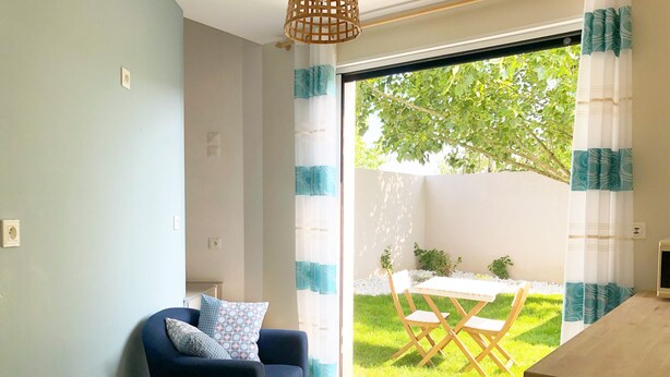 A modern living room with a sofa, a small table with a kettle and glasses, and a view of a garden through a sliding glass door. - Estrella (Les Sables-d'Olonne)