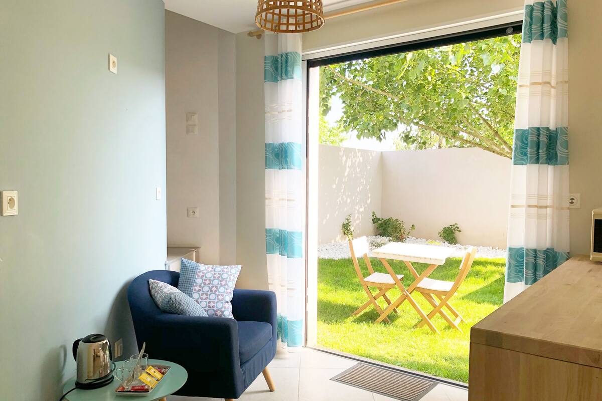 Una sala de estar moderna con un sofá, una mesita con una tetera y vasos, y vistas a un jardín a través de una puerta corrediza de vidrio.