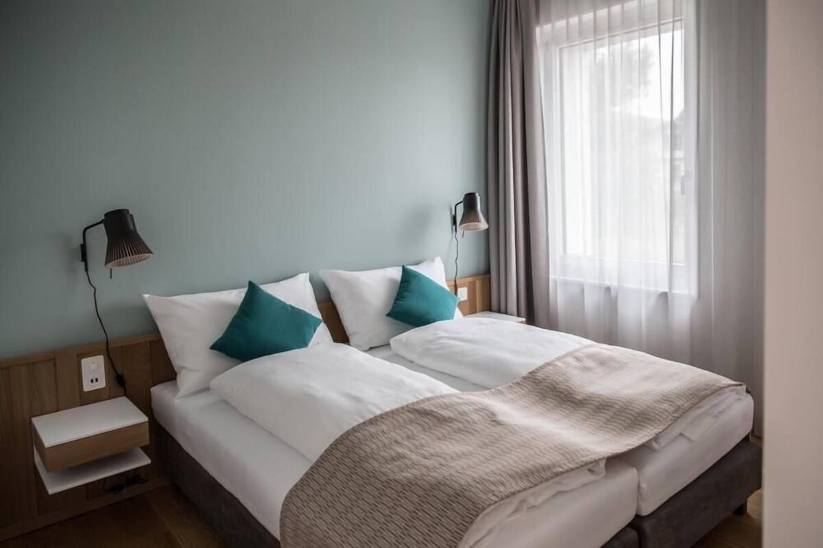 A neatly made double bed with white and teal pillows, a beige throw blanket, and two bedside lamps.