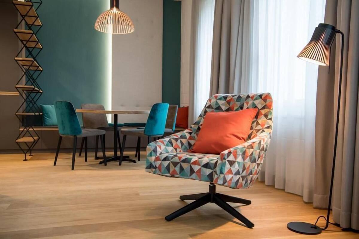 A modern living room with a geometric-patterned armchair, a dining table with chairs, a floor lamp, and a decorative wall piece.