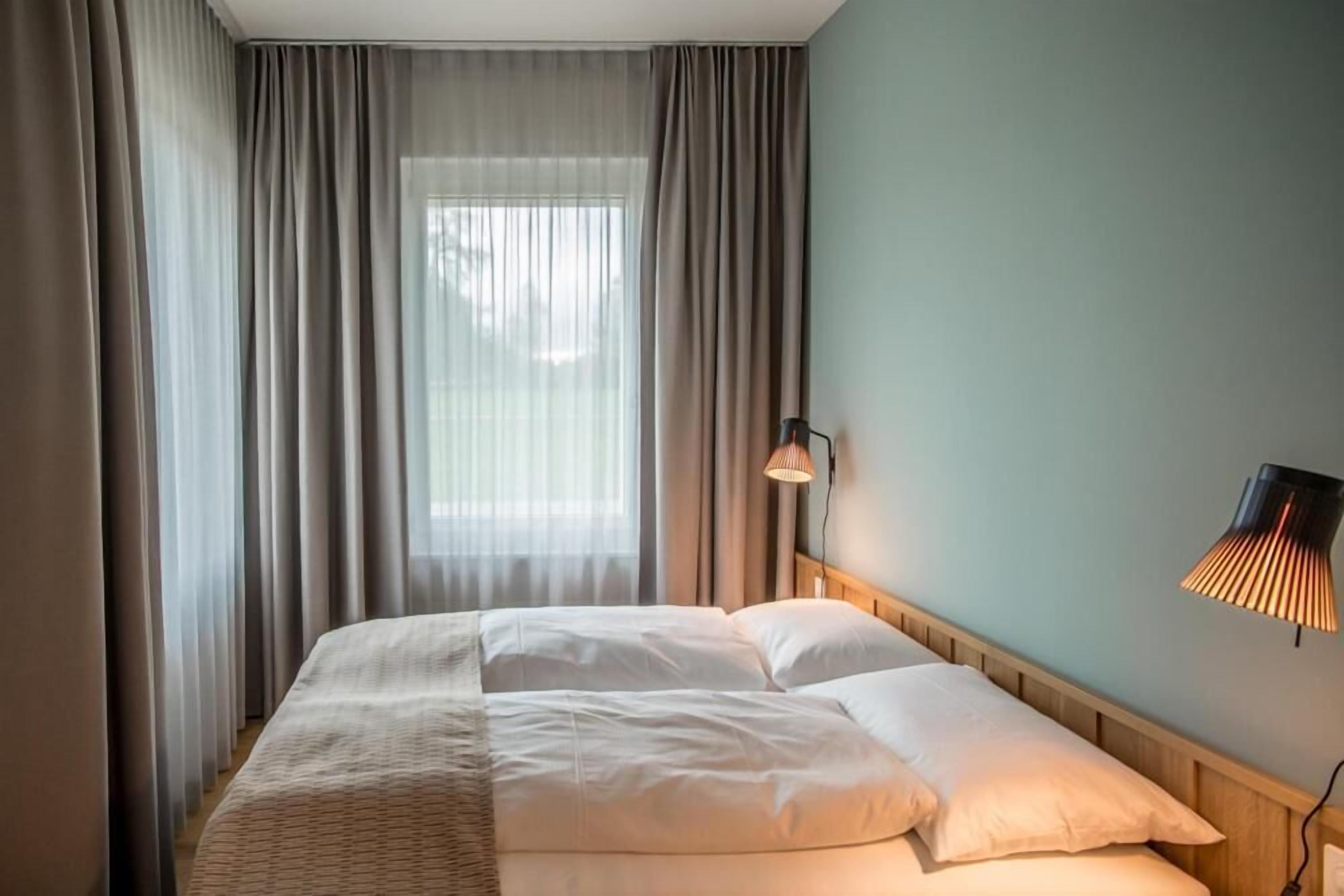 A hotel room with a bed, curtains, a wall-mounted lamp, and a window.