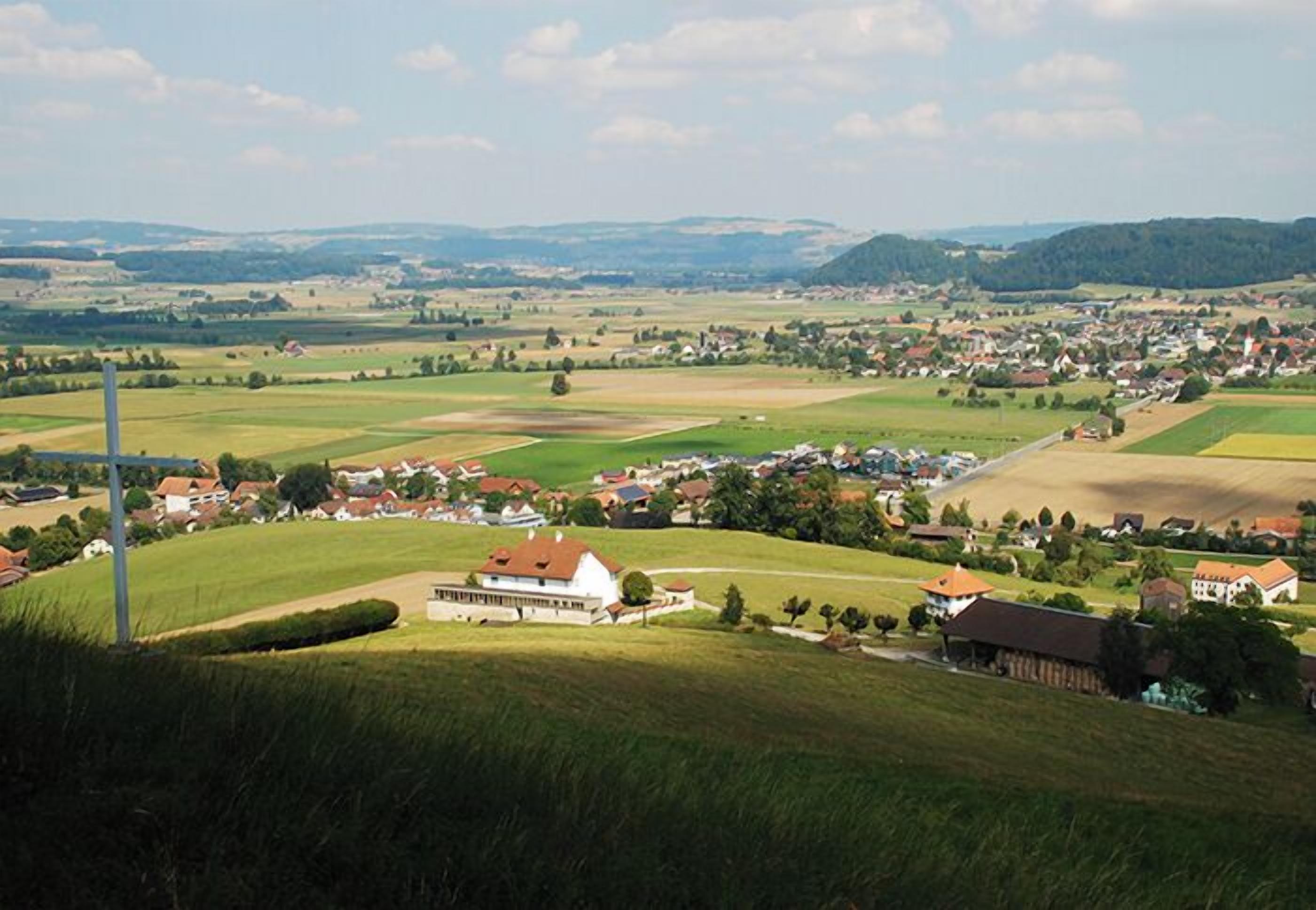 Un paysage rural dominé par un grand bâtiment blanc coiffé d’un toit rouge, entouré de champs et d’un petit village.