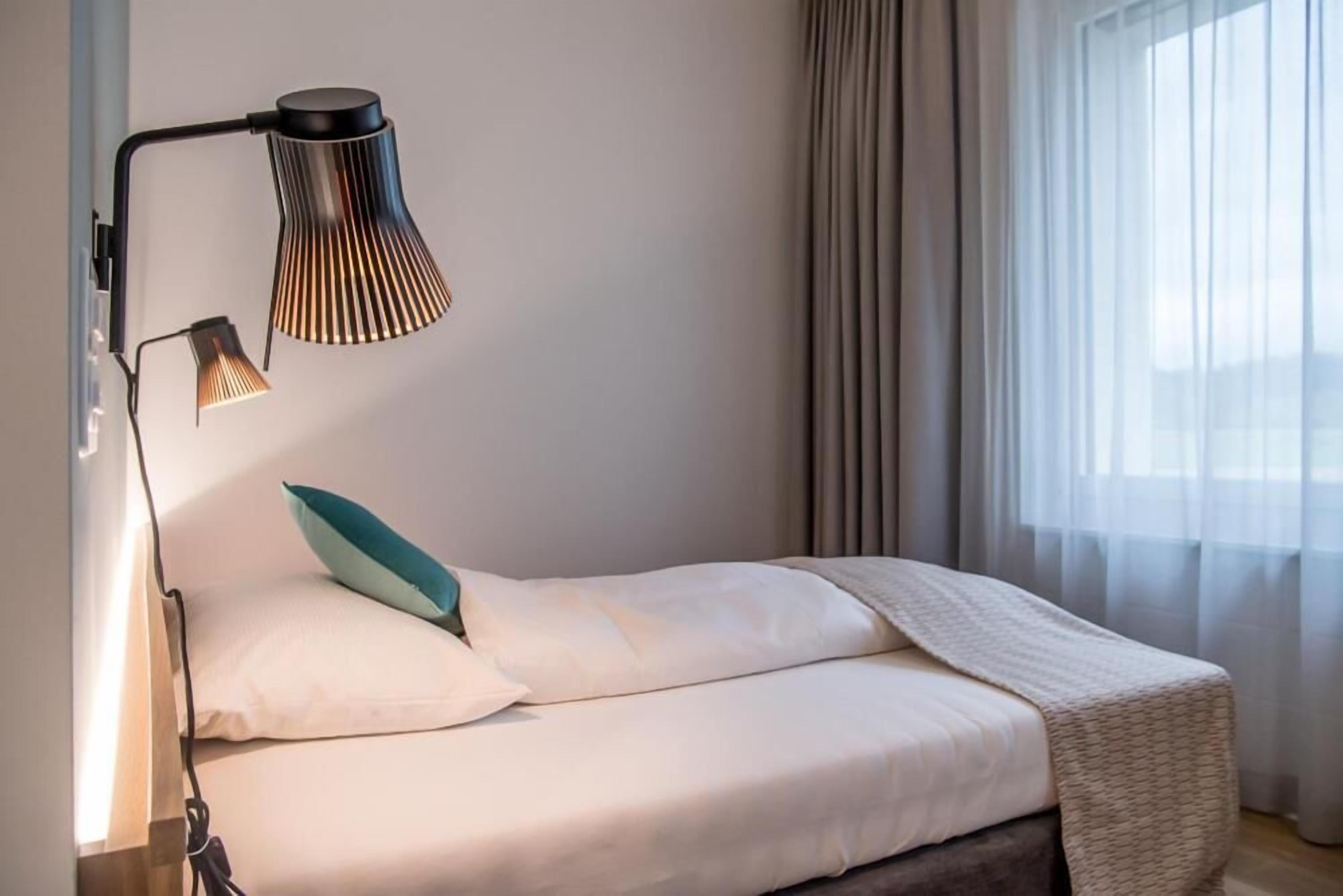 A neatly made bed with a white comforter and a teal pillow, a black adjustable desk lamp, and a window with sheer curtains.