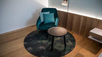 Ein Zimmer mit einem türkisfarbenen Sessel, einem runden Tisch, einem gemusterten Teppich, einer Stehlampe und einer Holzbank.