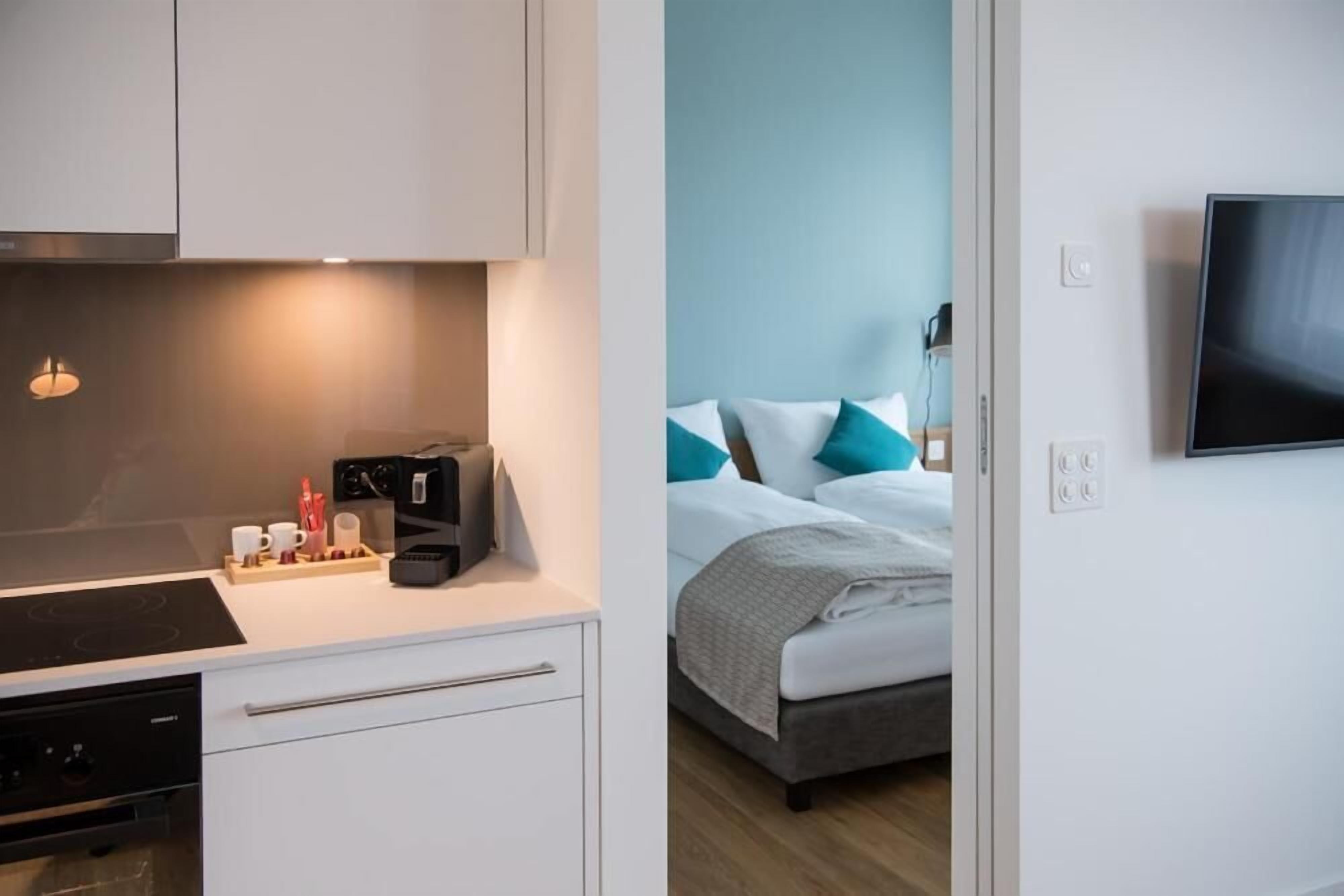 Une chambre d’hôtel moderne dotée d’une kitchenette, d’un lit avec une literie bleue et blanche, et d’un téléviseur à écran plat fixé au mur.