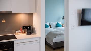 A modern hotel room with a kitchenette, a bed with blue and white bedding, and a flat-screen TV mounted on the wall.