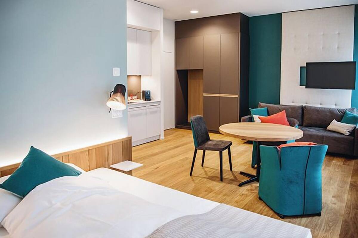 A modern hotel room with a bed, a round table, a sofa, and a TV mounted on the wall.