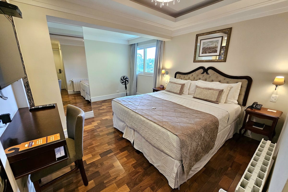 Una habitación de hotel con una cama grande, un escritorio con computadora, una silla, una ventana con cortinas y una mesita de noche con lámpara.