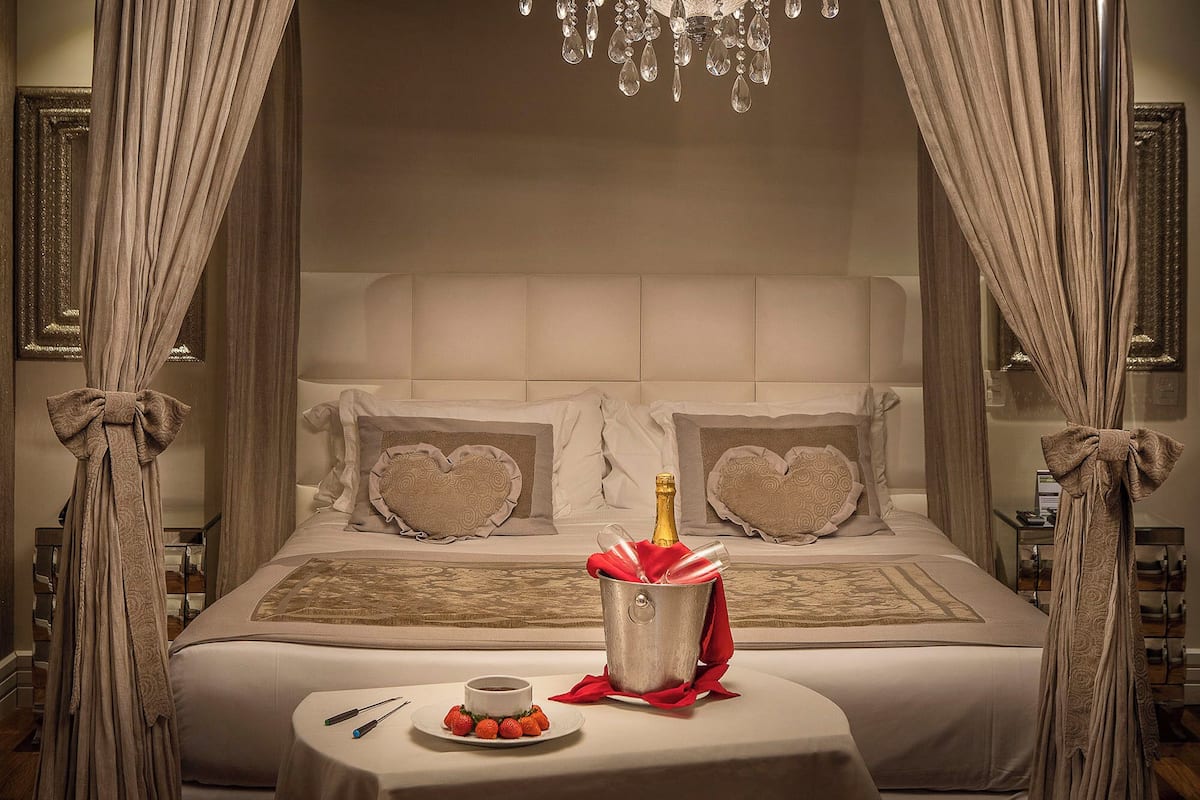 Un dormitorio lujoso con cama con dosel, almohadas con forma de corazón, una botella de champán y una bandeja de fresas.