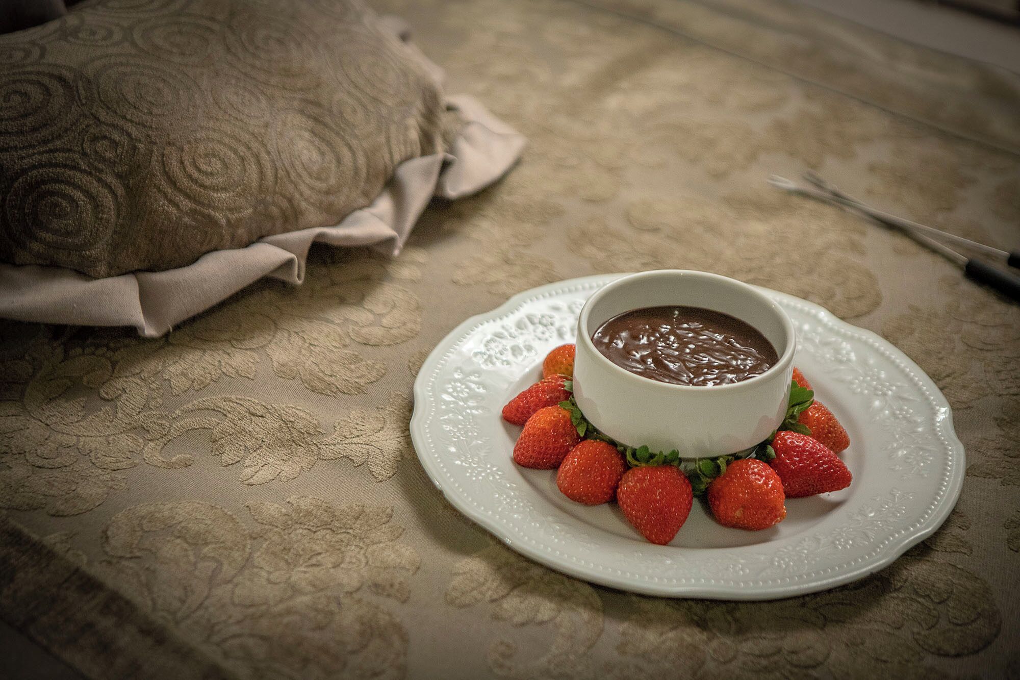 A bowl of chocolate soup on a white plate surrounded by strawberries, placed on a patterned tablecloth.