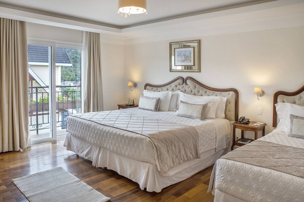 Un dormitorio con una cama grande, dos mesitas de noche, una mesita auxiliar, una ventana con cortinas y vistas al balcón.