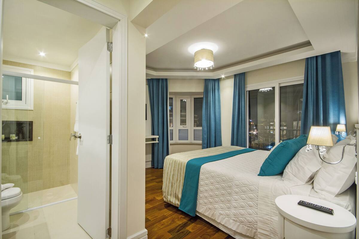 Habitación de hotel con cama, mesita de noche con control remoto, baño con cabina de ducha de vidrio y vistas a la ciudad a través de amplias ventanas.