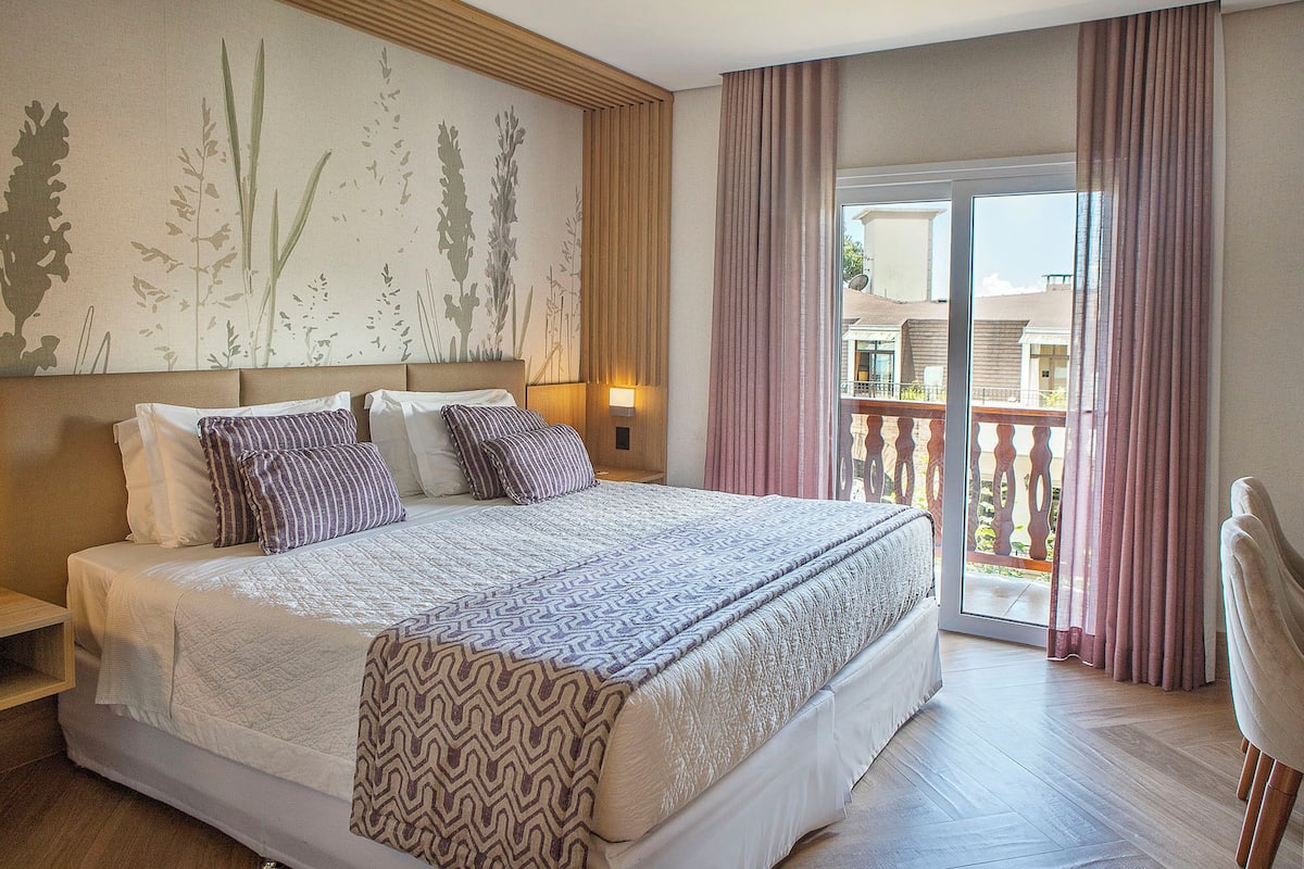 Habitación de hotel con una cama grande, una mesita de noche, una lámpara fijada a la pared y vistas al balcón exterior.