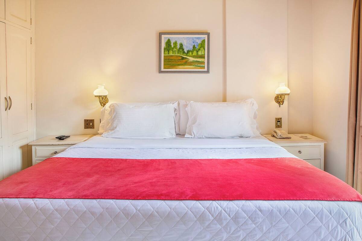 Habitación de hotel con cama, dos mesitas de noche, un cuadro en la pared y una colcha roja sobre la cama.