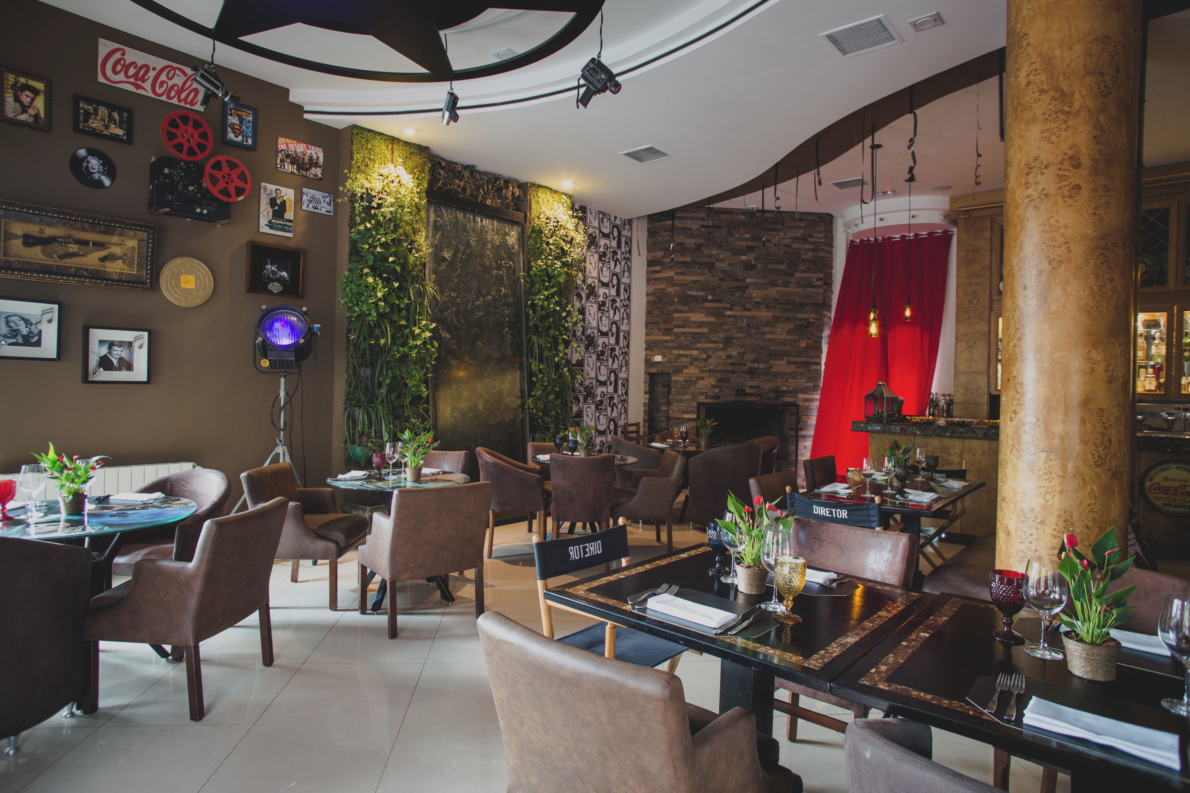 A restaurant with multiple dining tables and chairs, a wall with framed pictures and a Coca-Cola sign, and a red curtain in the background.