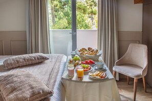 Habitación de hotel con cama, una mesa de desayuno con alimentos, una silla y vistas a la vegetación por la ventana.