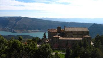 Edificio histórico de piedra con tejado de tejas, rodeado de abundante vegetación y con vistas a un valle con un lago.