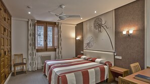 A hotel room with a large bed, a desk, a chair, a window with curtains, a ceiling fan, and wall art. - Hotel Monasterio de Leyre (Yesa)