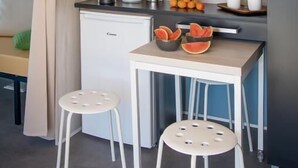 Une cuisine compacte avec un plan de travail blanc, deux tabourets, un petit réfrigérateur et une plante posée sur l’étagère.