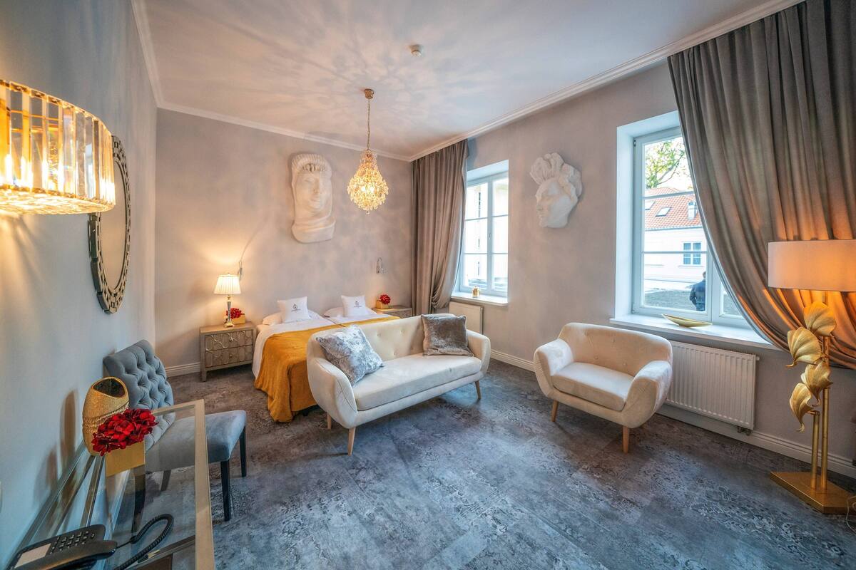 Une chambre d’hôtel avec un lit, deux fauteuils, une table en verre avec un téléphone, une lampe et une grande fenêtre avec des rideaux.
